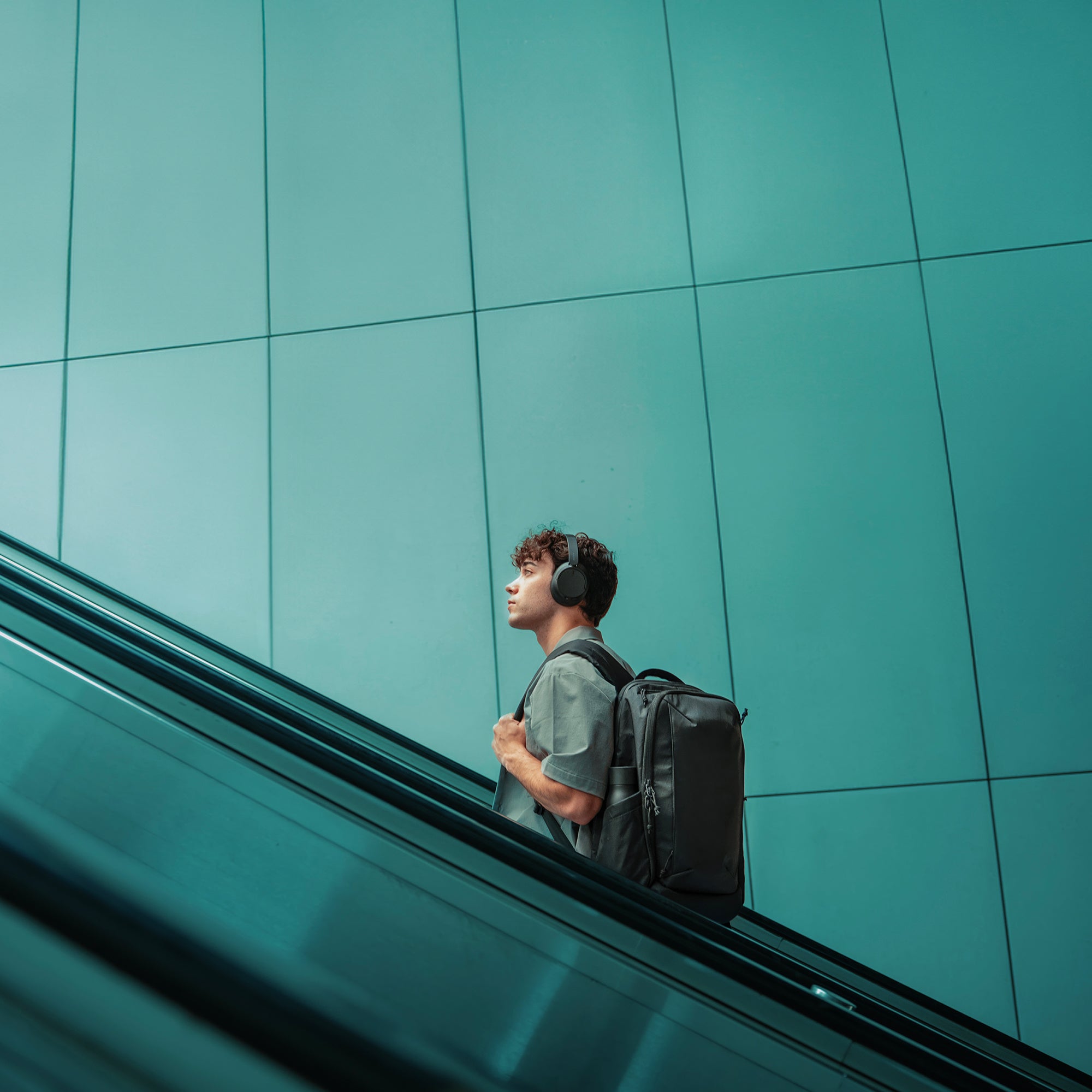Person with a backpack and headphones standing on an escalator against a teal wall.